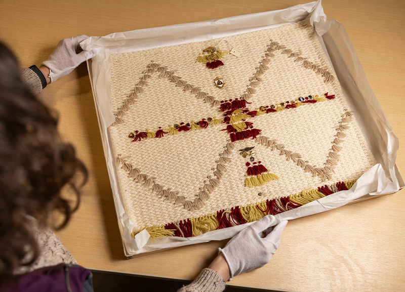 Gloved hands present a woven textile with red and tan patterns, pinned with small vintage emblems