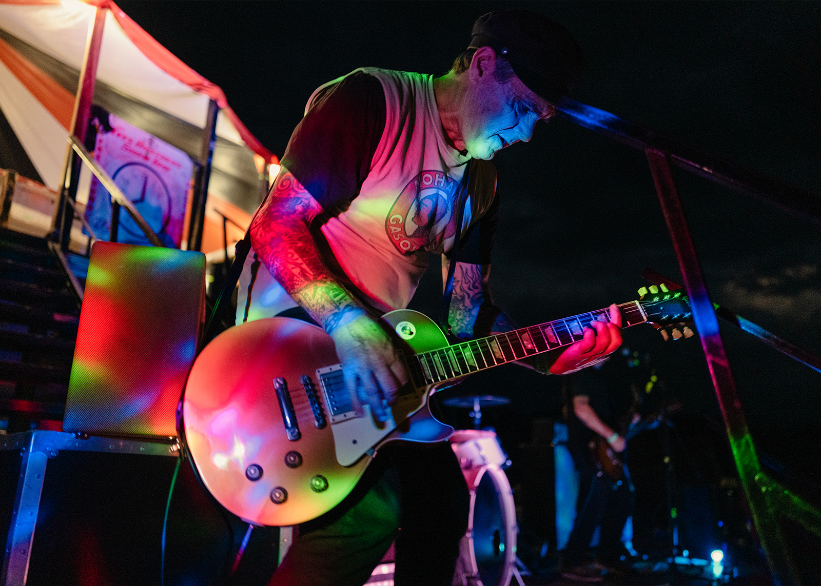 A tattooed guitarist playing a Les Paul electric guitar performs under colorful lights at night.