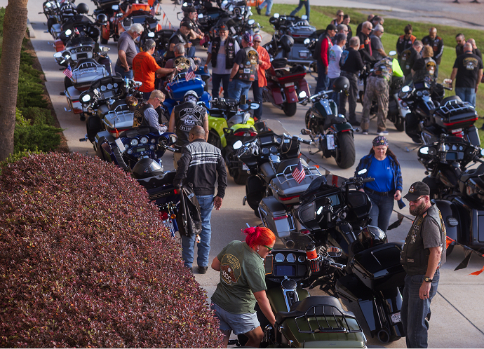 Rows of parked touring motorcycles line a sidewalk as riders gather and walk between bikes at an event