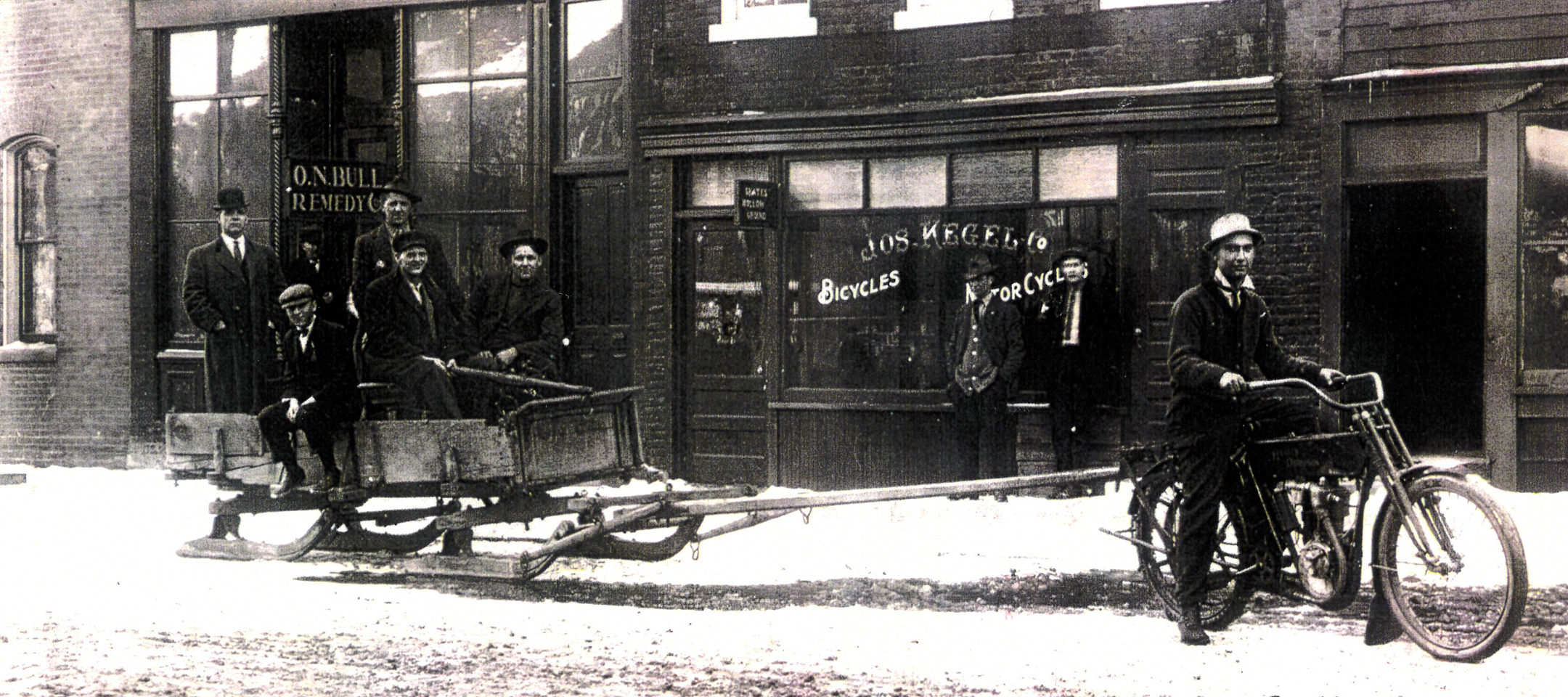 A Photo from 1909 of a motorcycle pulling a wooden sled on a snowy street outside of Joe Kegel’s Bicycles & Motorcycles