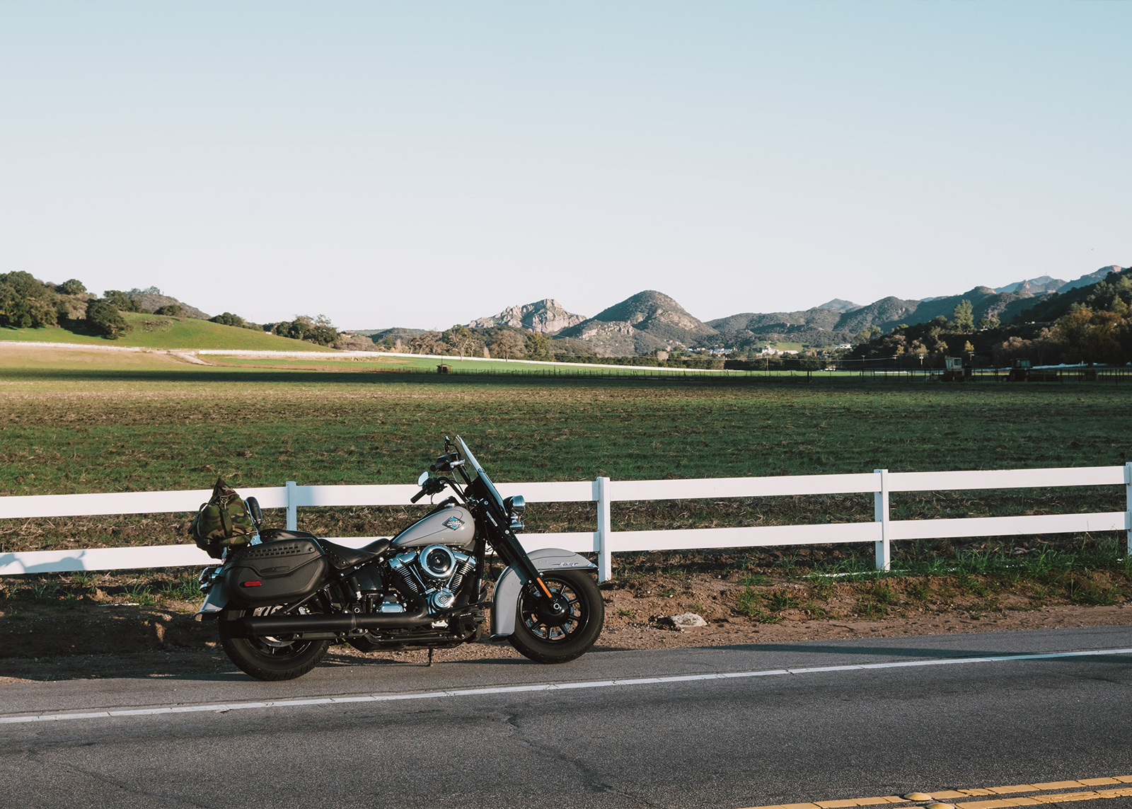 A Harley-Davidson Heritage Classic motorcycle parked next to a white fence with a farm and mountains in the background