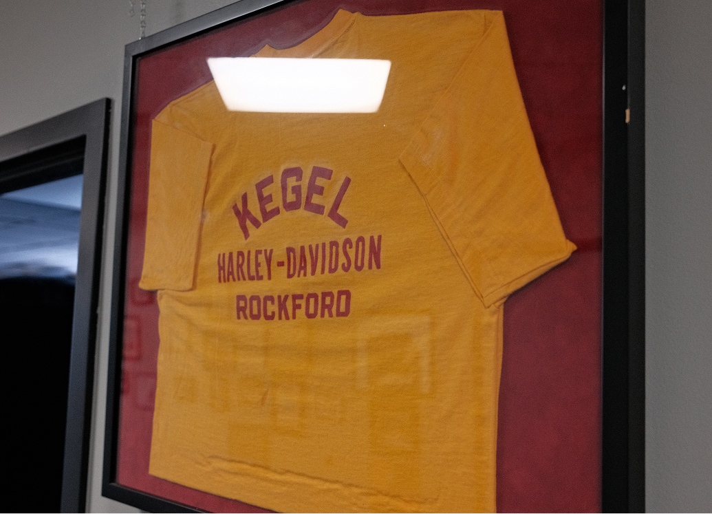 A vintage yellow shirt with red letters that say Kegel Harley-Davidson, Rockford is mounted and hung in a frame
