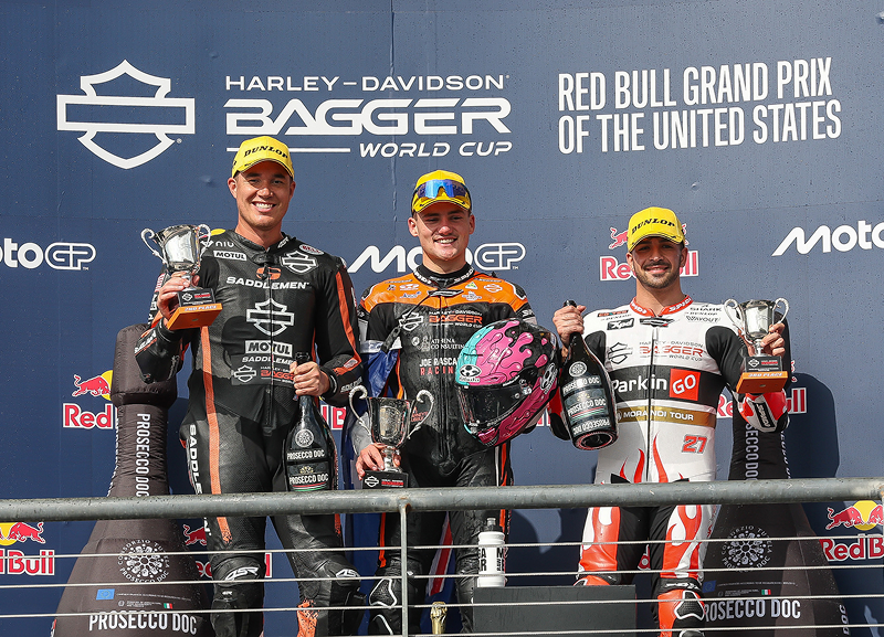 Three riders stand on a podium holding trophies beneath Harley-Davidson Bagger World Cup signage