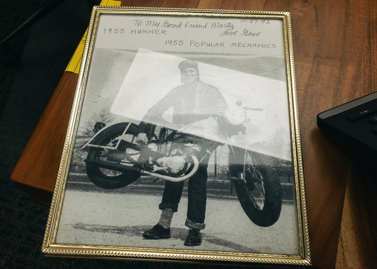 A framed vintage photo of a man lifting a motorcycle off the ground with a personal message handwritten across the top.