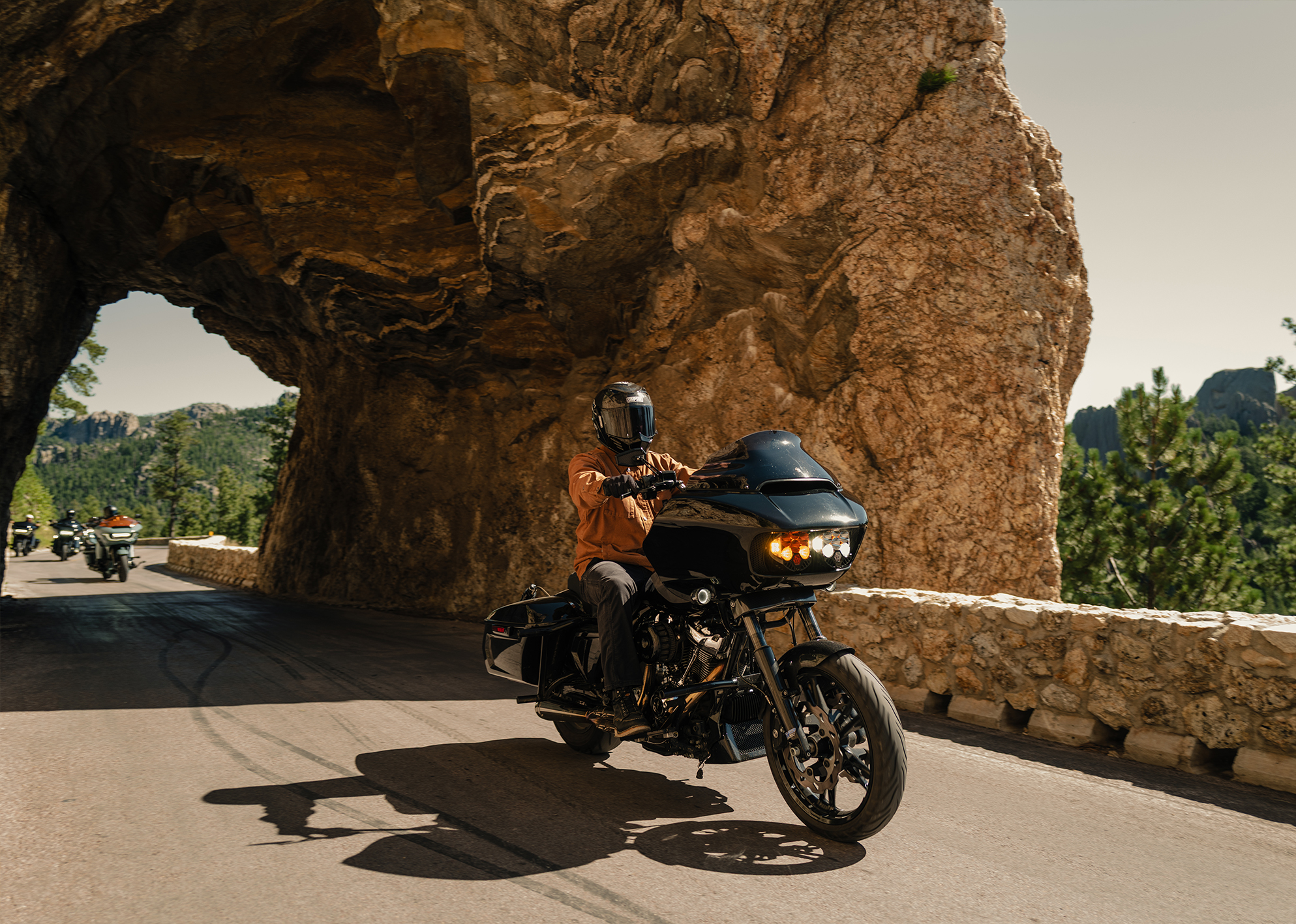 Motorcyclist rides through a scenic rock tunnel with other riders in the background.