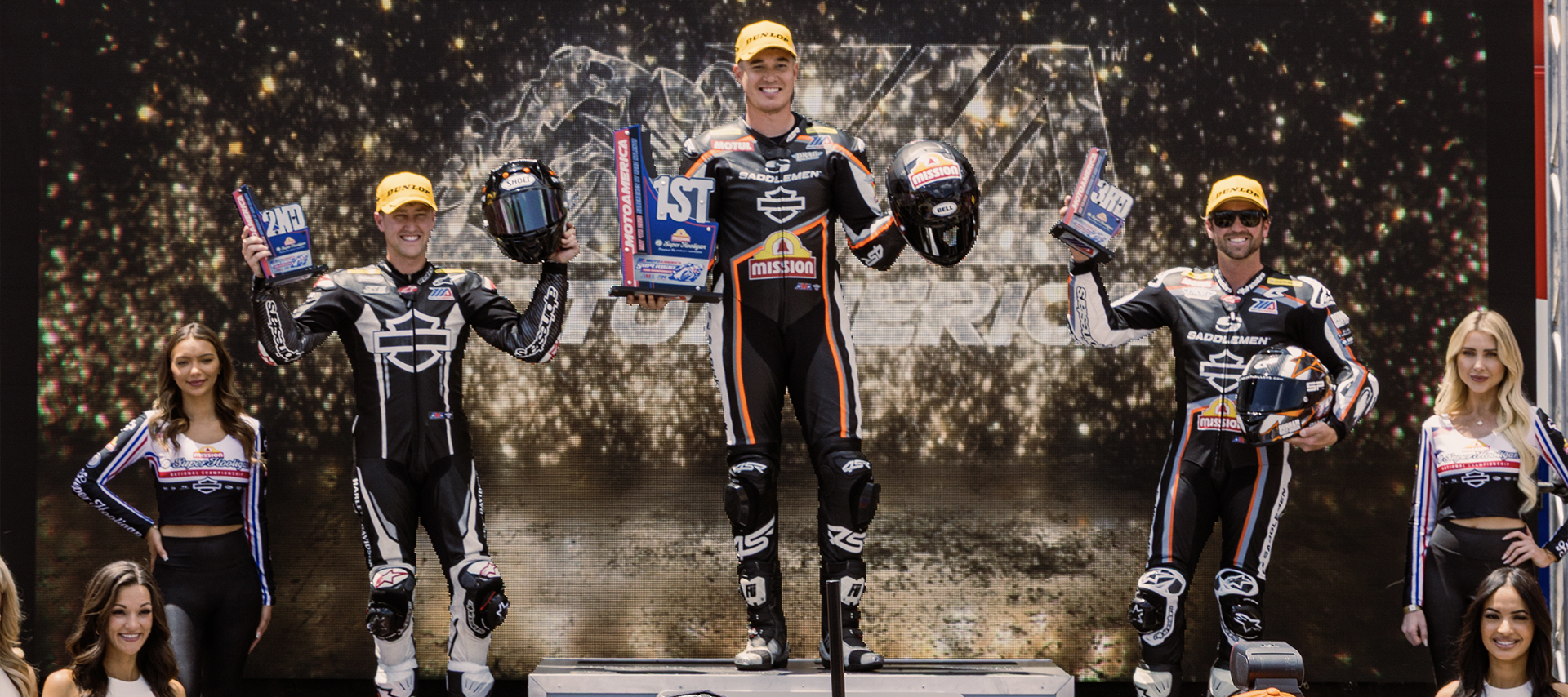 HD racing team members stand on the podium holding 1st, 2nd, and 3rd place trophies. Infront of them are a crowd of cameras capturing the moment.  