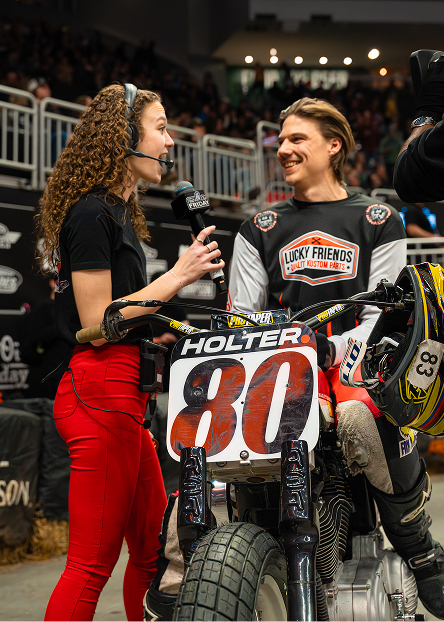 A broadcaster interviews a racer beside a flat‑track motorcycle in the pit area before a race