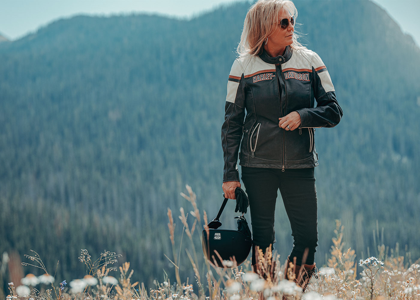 Woman in a field wearing a H-D riding jacket and carrying her helmet