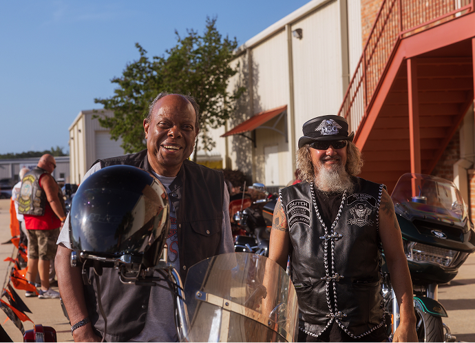 Two riders stand beside parked motorcycles outside a building, wearing vests and helmets at an event