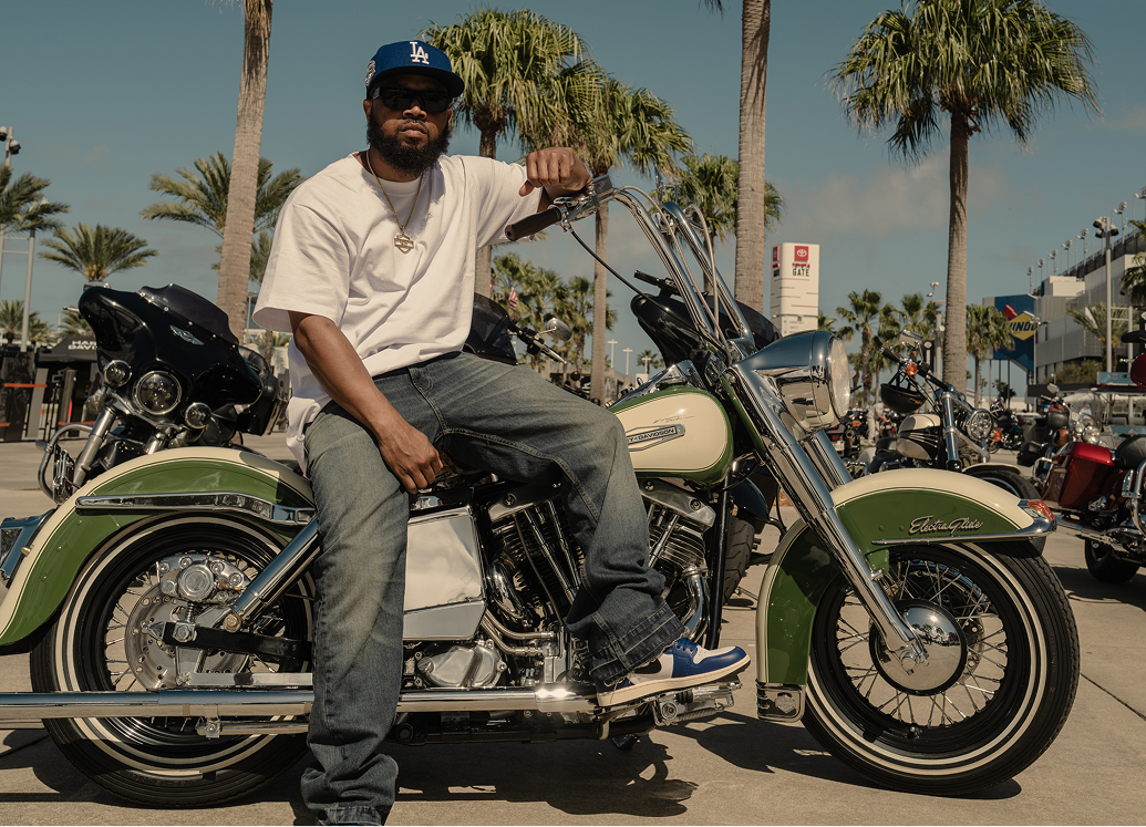 Rider seated on a classic green motorcycle in a sunlit parking area filled with other bikes