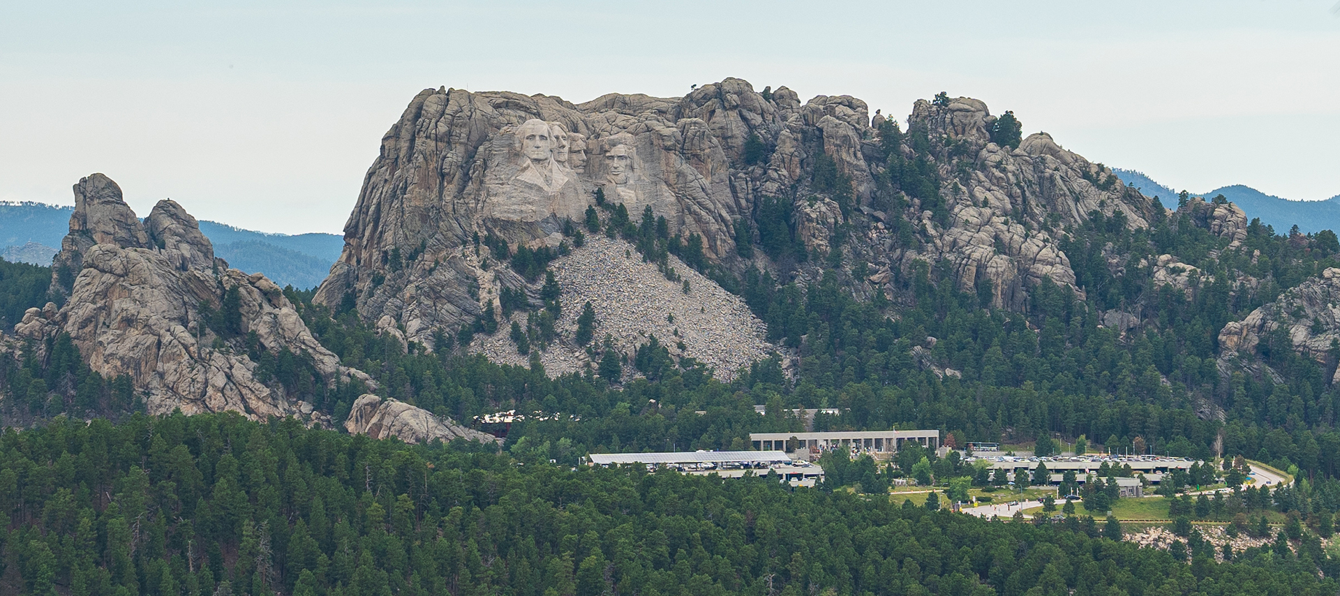Distant view of Mount Rushmore framed by pine trees, with forested hills and visitor facilities below.