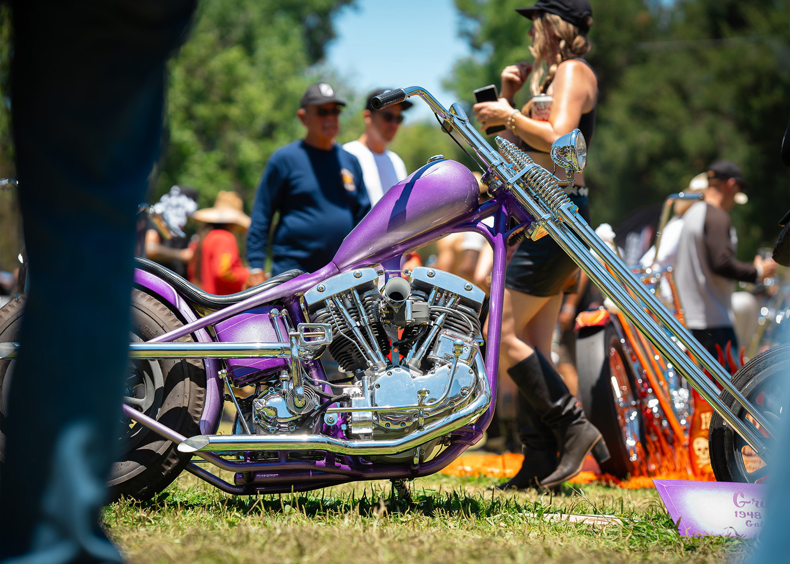 A purple and chrome chopper parked on the grass with a woman in leather boots and top standing behind it.