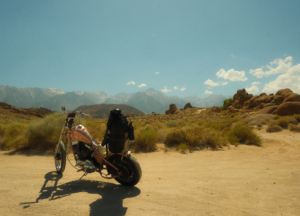 Custom chopper motorcycle parked on a dirt road with desert terrain, boulders, and mountain range beyond