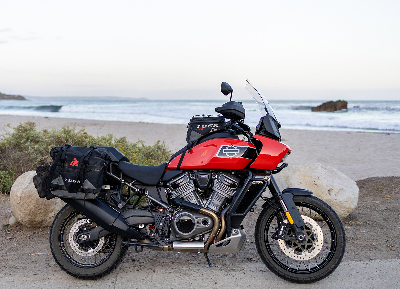 Red H-D Pan America motorcycle with side bags parked on sandy beach overlook with waves and rocks offshore