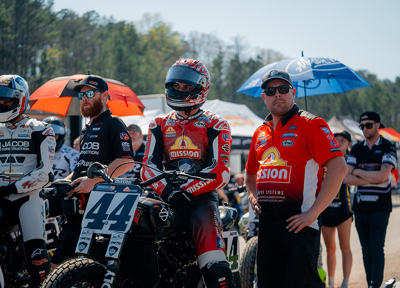 Number 44 Brandon Robinson waits on grid beside crew under umbrellas with riders lined up behind
