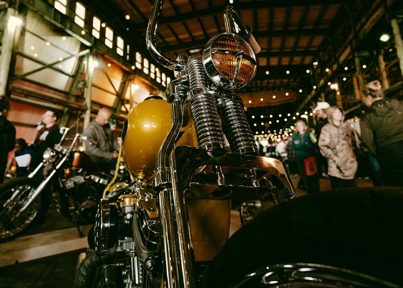 A closeup of a bike, looking up at a yellow tank and amber headlight, and chrome forks among a crowd of people