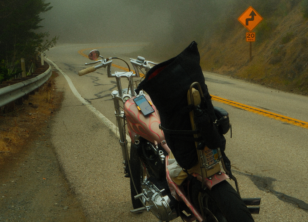 Custom chopper motorcycle loaded with travel gear parked on a winding coastal highway in foggy conditions