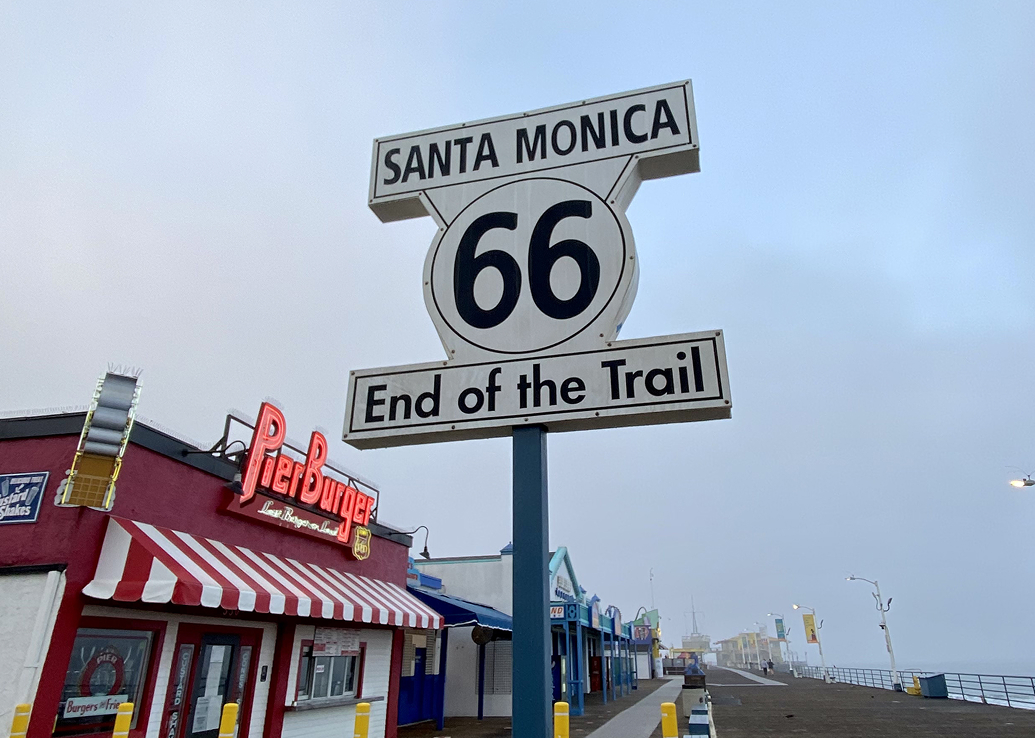 Route 66 ‘End of the Trail’ sign on the Santa Monica Pier, with shops and the boardwalk visible