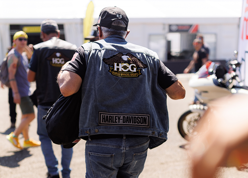 H.O.G. member in a Harley-Davidson vest walks through a motorcycle event with riders and bikes nearby
