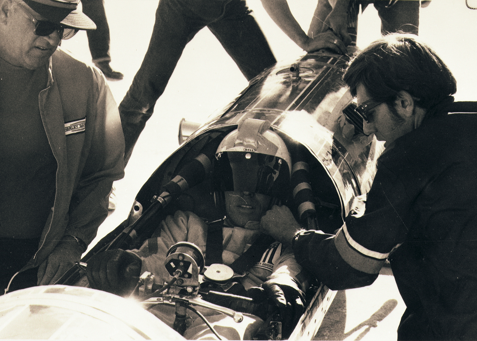 One man sits inside the streamliner, strapped in and helmet on, while two other men stand around him checking his straps.