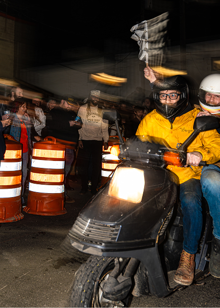 Two riders on a scooter move through a crowd, one raising a checkered flag while passing traffic barrels