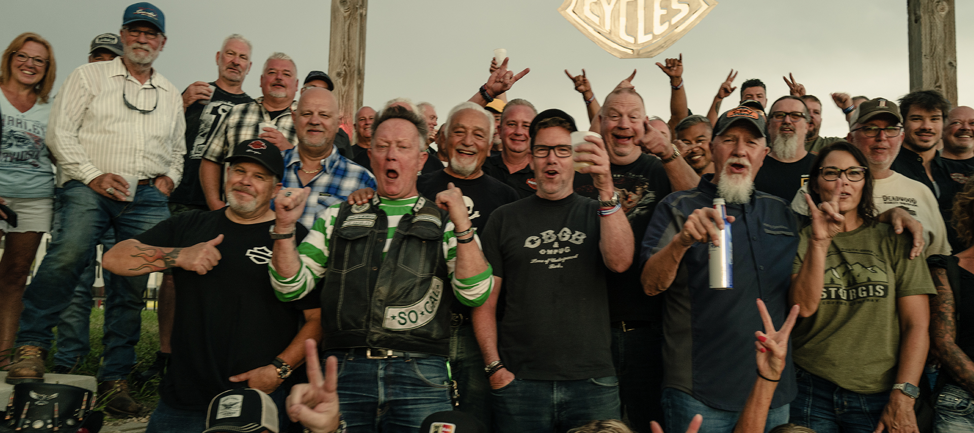 Large group of bikers pose together, smiling and cheering during a Sturgis rally gathering.
