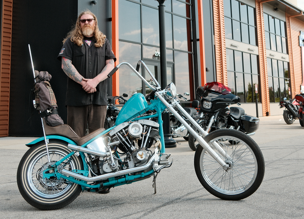 Custom turquoise Harley-Davidson chopper parked outside dealership with rider standing beside it