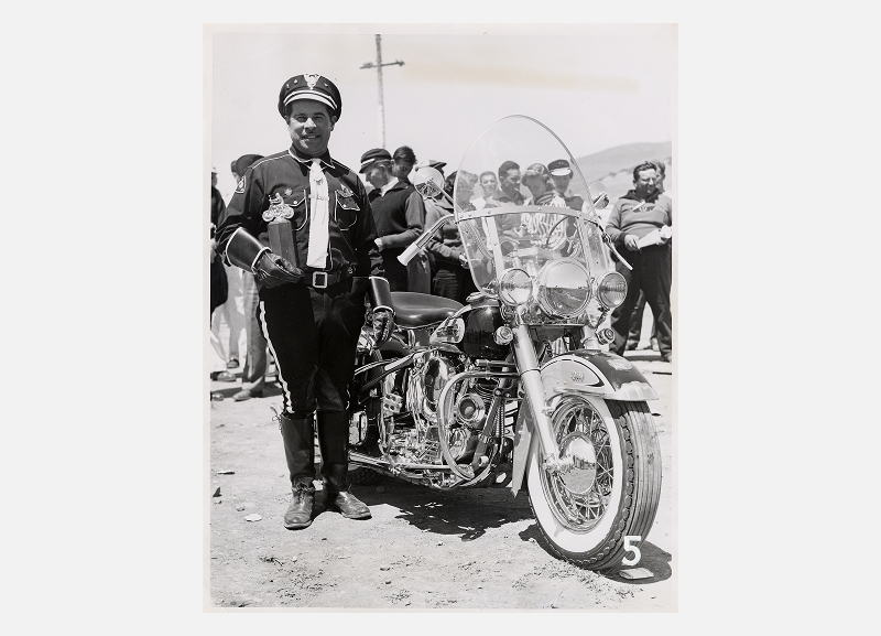 Vintage portrait of a motorcycle rider in uniform standing beside a Harley-Davidson at a gathering
