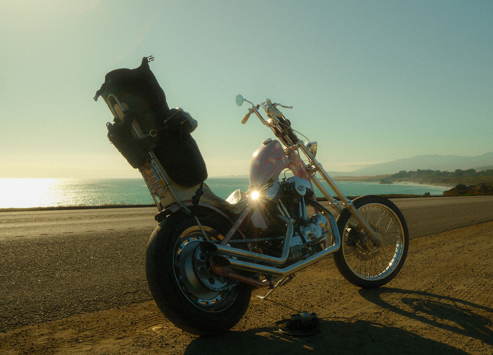Custom chopper motorcycle parked along a coastal highway at sunset, with ocean views, rugged shoreline, and travel gear loaded on the bike