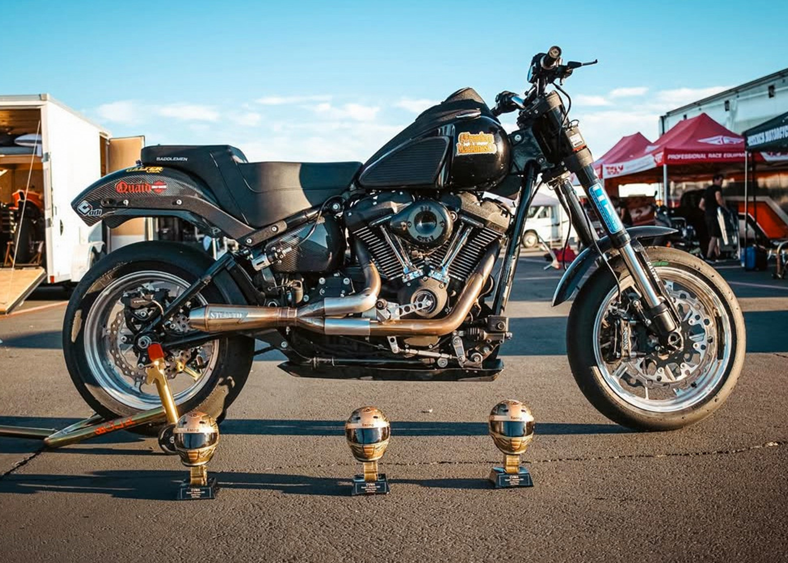 A side angle of one of Quaid’s racing motorcycles with three trophies displayed on the ground in front of it.