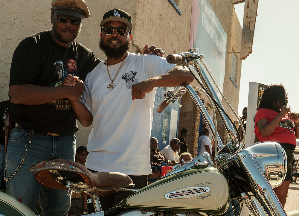 Two people clasping hands and posing beside a vintage-style motorcycle on a neighborhood street with other riders nearby