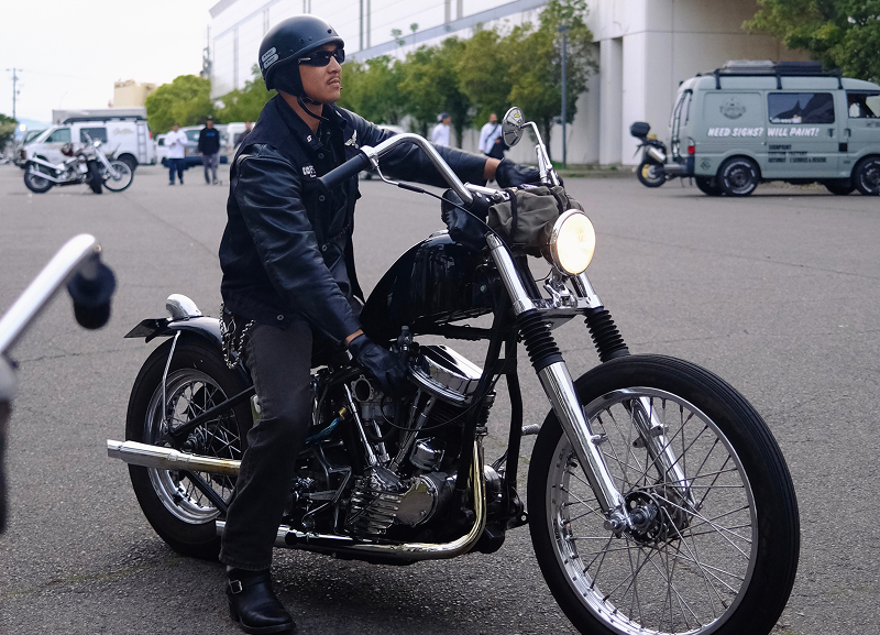 Rider sitting on a dark custom motorcycle parked on pavement with industrial buildings and cars in background