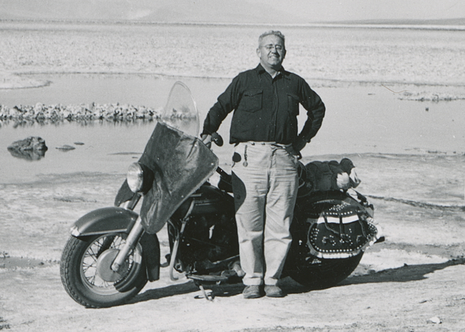  Black and white historical photo of Eldred Harrington standing by his 1951 Harley-Davidson Hydra Glide motorcycle in the desert.