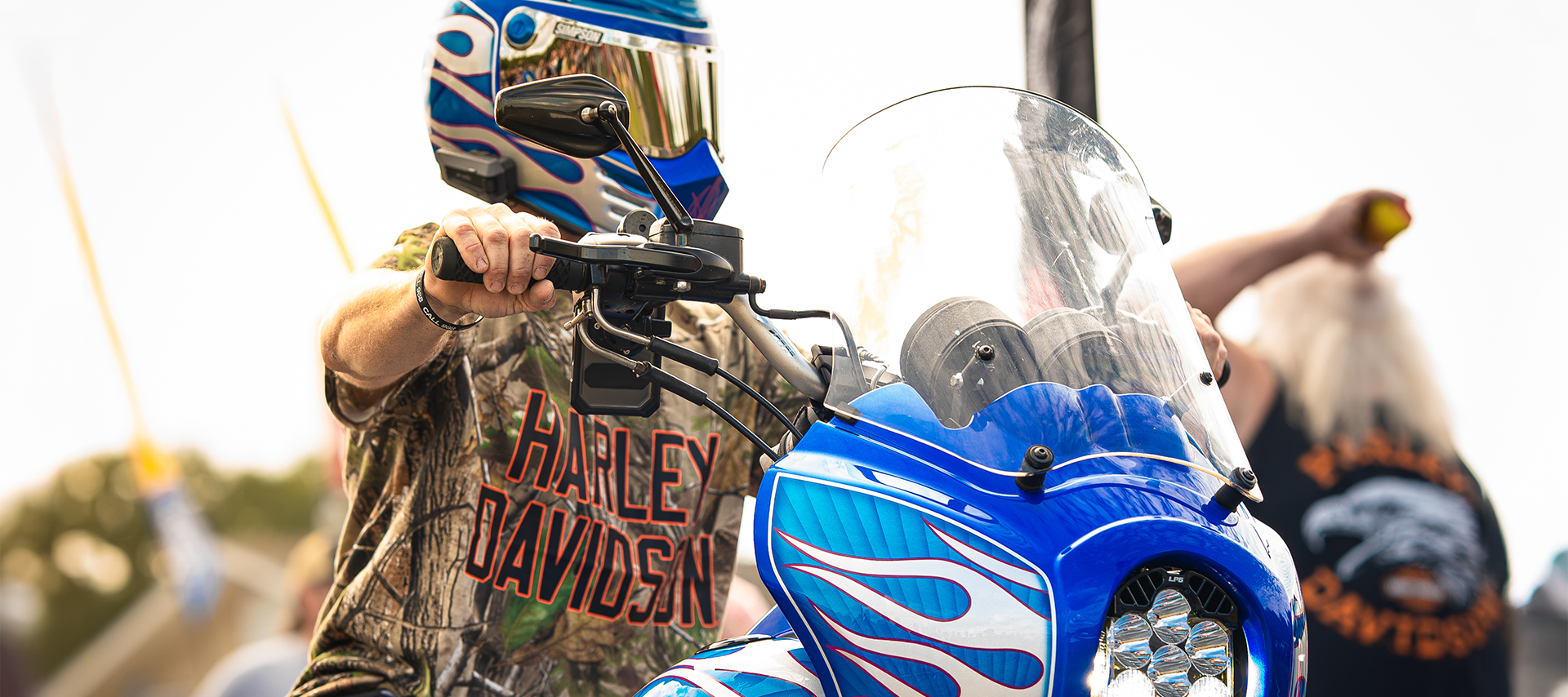 Rider in Harley-Davidson X Realtree shirt and blue flame helmet grips the handlebars of a custom motorcycle.