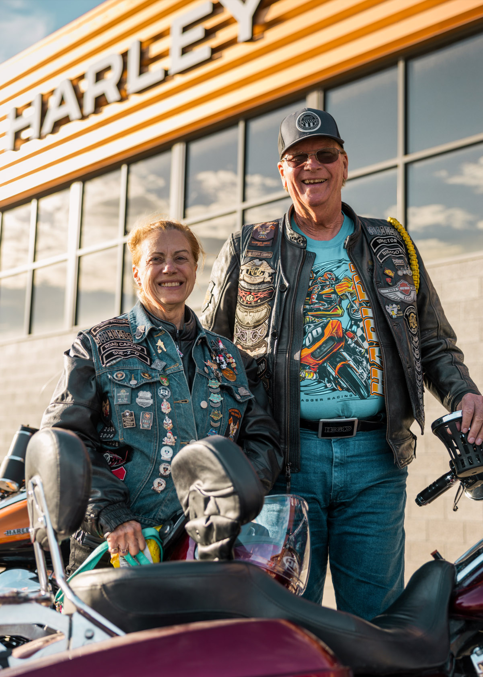 Pair of motorcycle enthusiasts with vests and pins standing by touring bikes at a Harley location