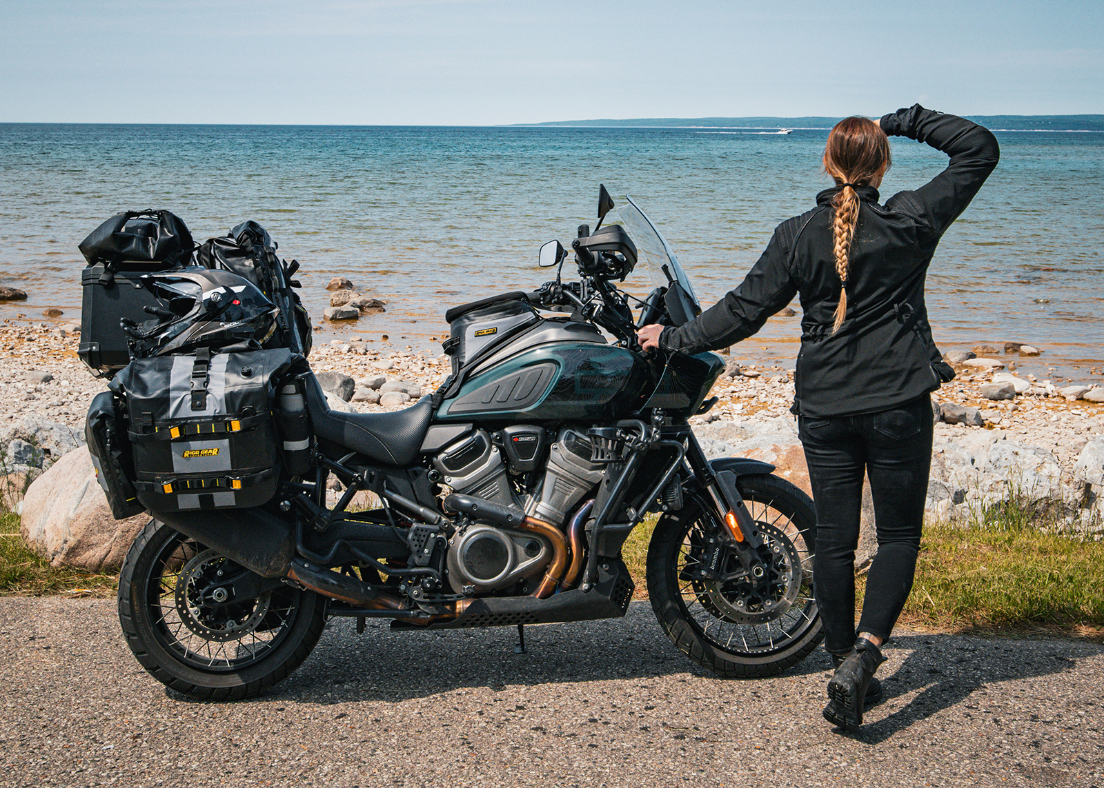 The author stands beside her packed Harley-Davidson Pan America motorcycle, looking out over a rocky shoreline.