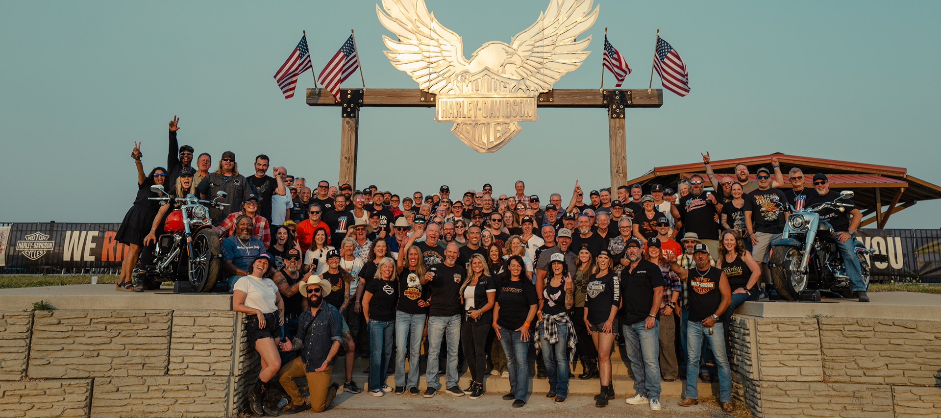 A group photo of the Harley-Davidson dealer network gathered beneath an iconic bar-and-shield sign during Sturgis Rally