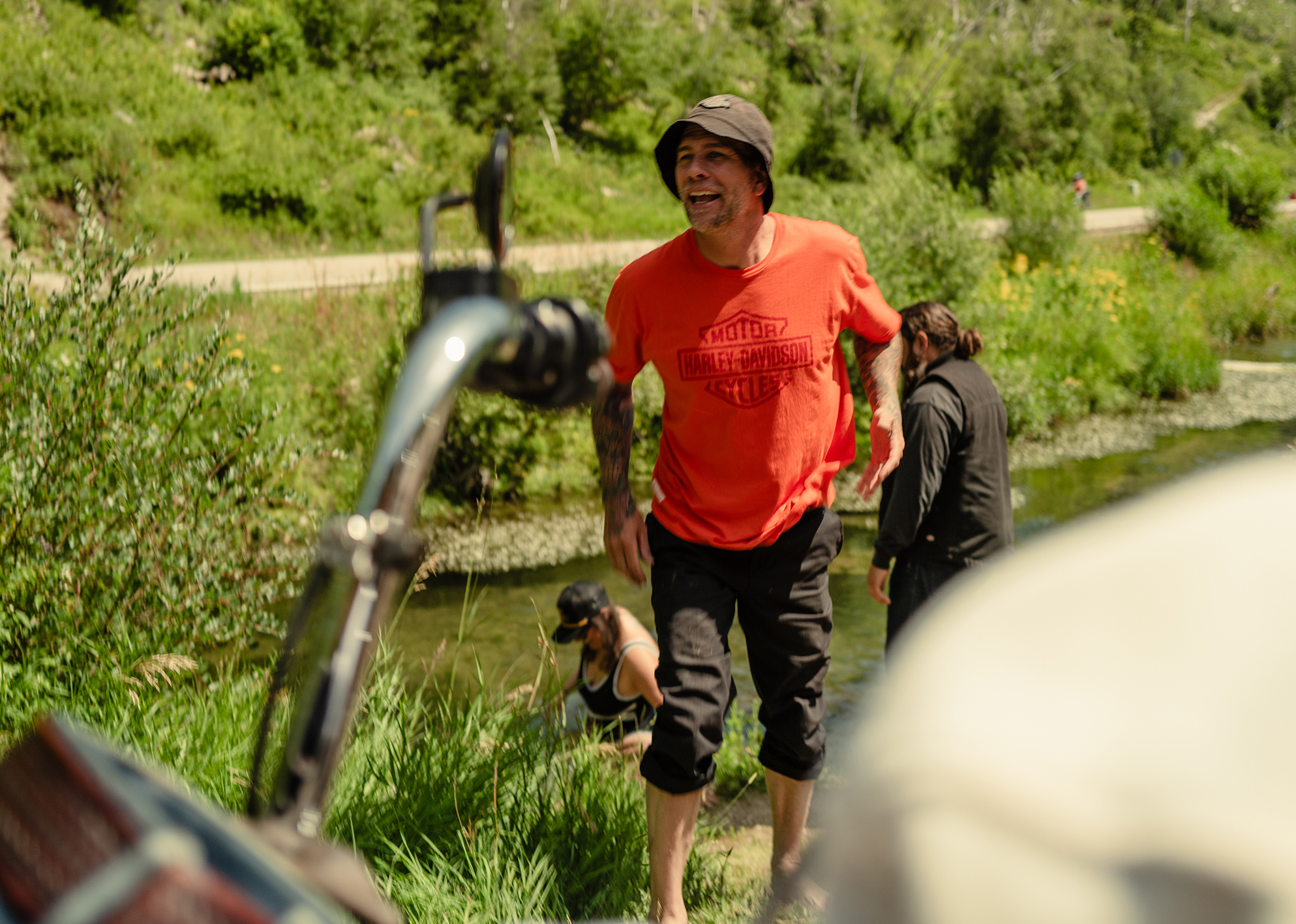 Man in orange Harley-Davidson shirt smiles while standing by a stream in a green mountain valley.