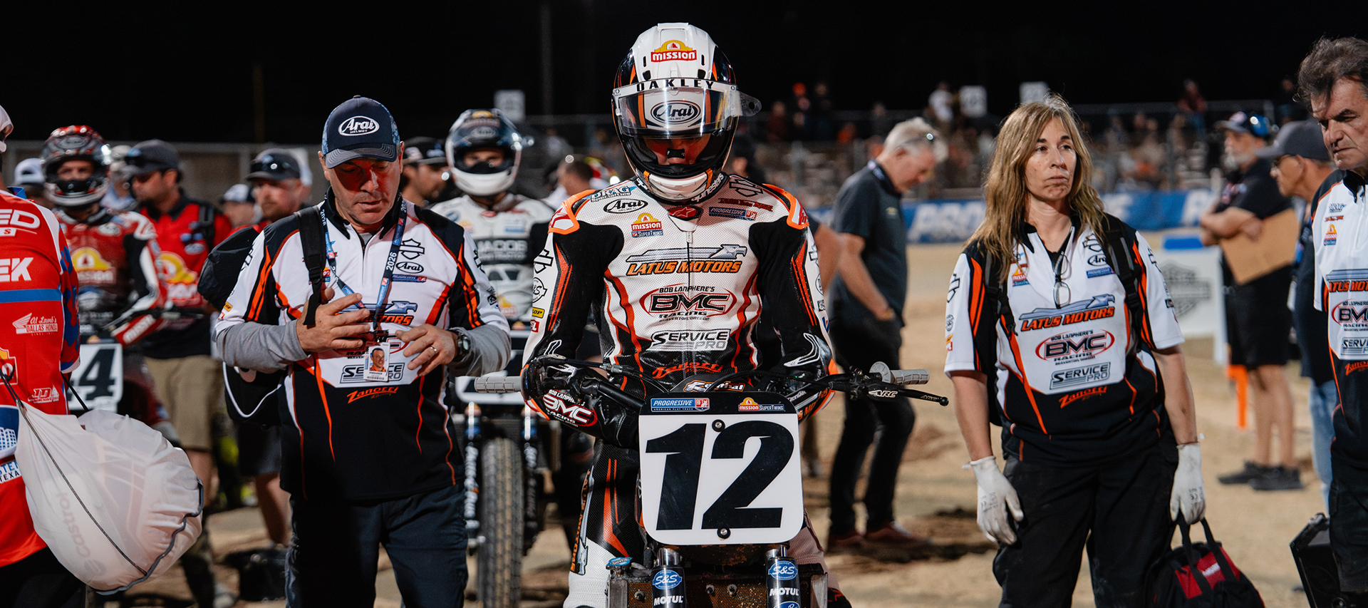Flat track racer Kody Kopp waits on the track before competition as crew members and riders gather under stadium lights