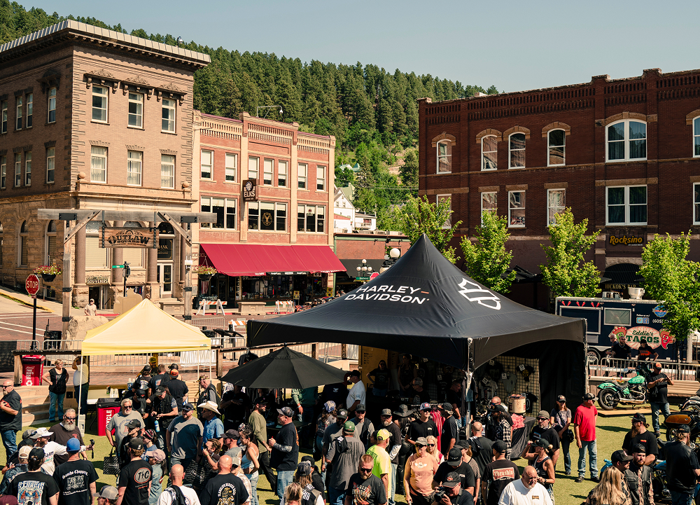 Crowd gathered around H-D tent in historic downtown with brick buildings and hills in background.