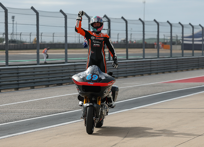 Race winner stands on his motorcycle with a raised fist while riding through pit lane after crossing the finish line