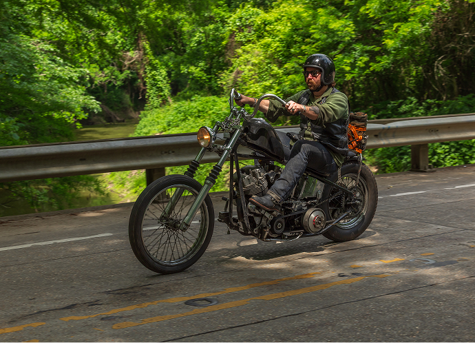 Payton Broussard crosses a bridge on his custom black motorcycle, surrounded by dense green trees along a winding road