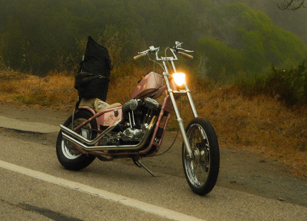 Custom chopper motorcycle parked along a coastal roadway with hillsides, fog, and illuminated headlight