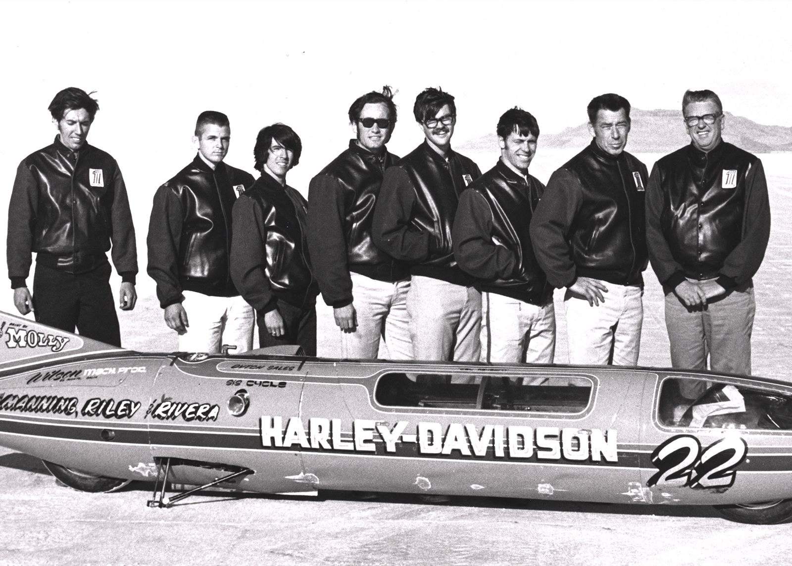 Eight men in black Harley jackets stand behind the Sportster streamliner in the desert. 