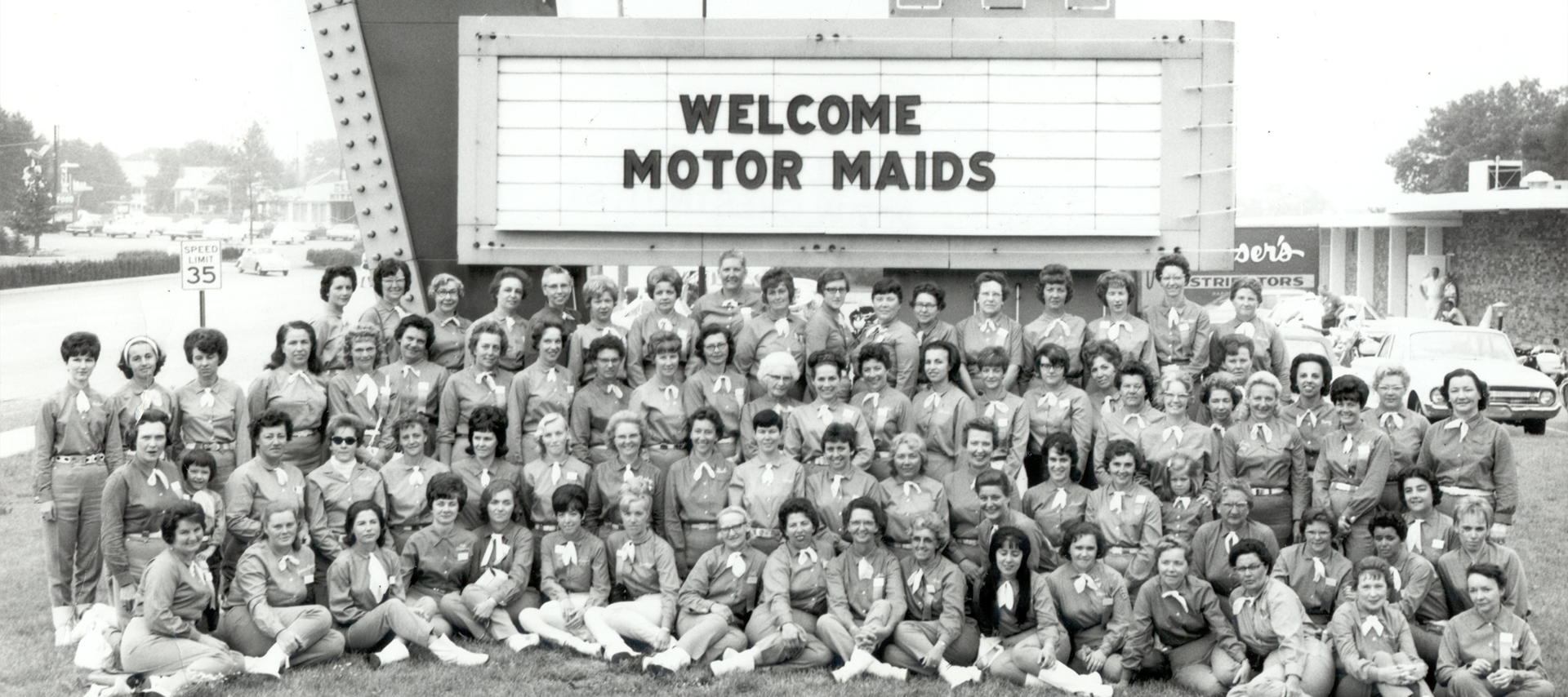 A vintage black and white group photo of the Motor Maids in front of a large Holiday Inn sign and marquee that says “Welcome Motor Maids” during a rally in 1967.
