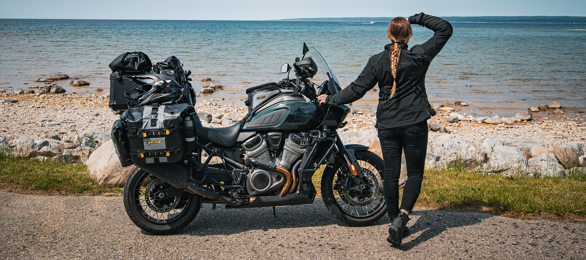 The author stands beside her packed Harley-Davidson Pan America motorcycle, looking out over a rocky shoreline.