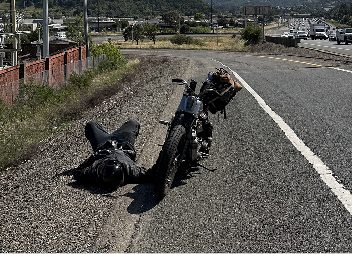 Motorcycle parked on a roadside shoulder with rider lying on the ground nearby beside a busy highway