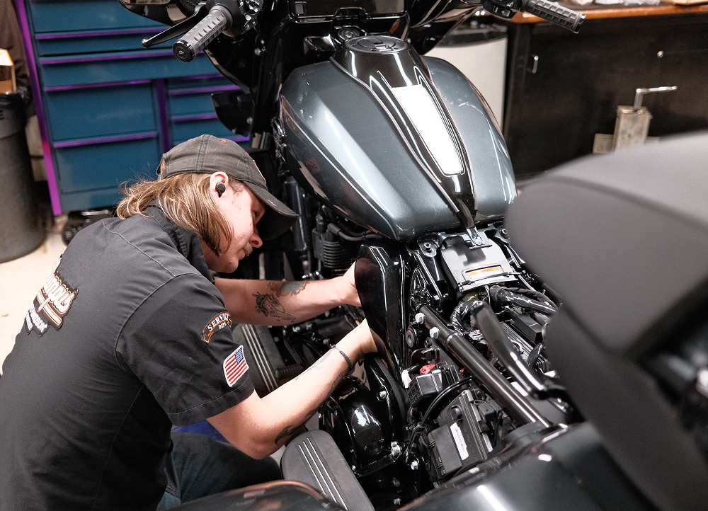 Technician working on a Harley-Davidson motorcycle engine inside a workshop with tool cabinets and service equipment