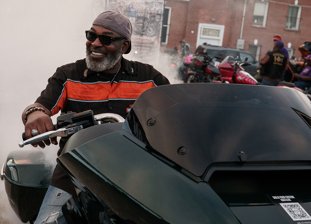 Rider seated on a large touring motorcycle with matte fairing, parked on a city street during a group ride