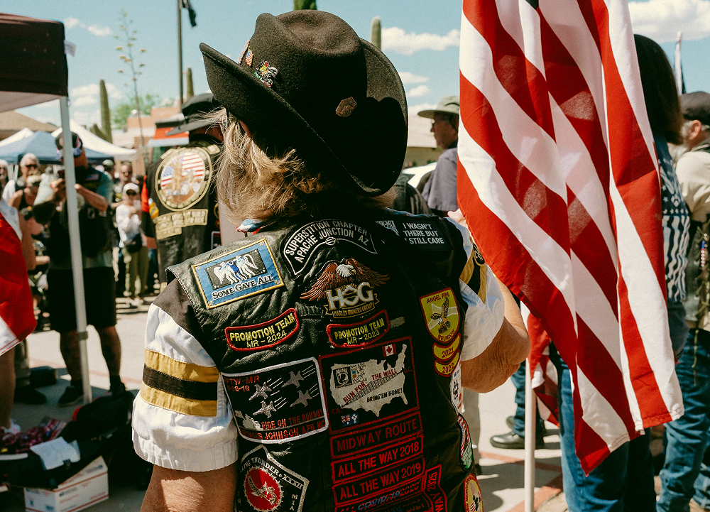 Motorcycle rider seen from behind wearing a vest covered in patches, standing near an American flag at a bike event